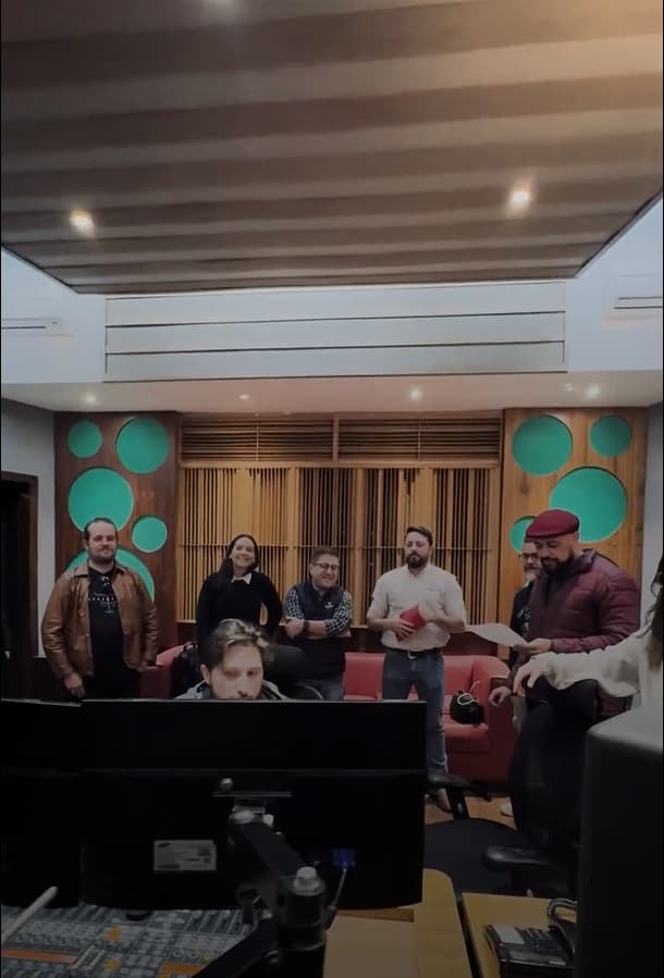 Interior de um estúdio de gravação com várias pessoas observando o trabalho por trás de uma mesa de som.