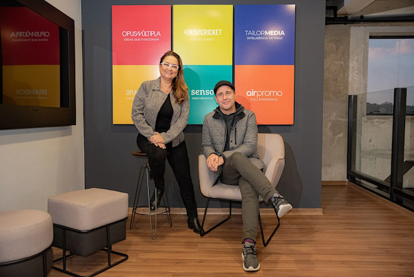 Ambiente interno do Grupo OM, em Curitiba, exibindo painéis coloridos com as marcas que compõem o ecossistema do grupo — incluindo HouseCricket, OpusMúltipla, TailorMedia, Brainbox, Airpromo e Sense. Dois profissionais posam sorridentes no espaço moderno, com decoração clean e iluminação natural, transmitindo o clima colaborativo e criativo que caracteriza a cultura da empresa.