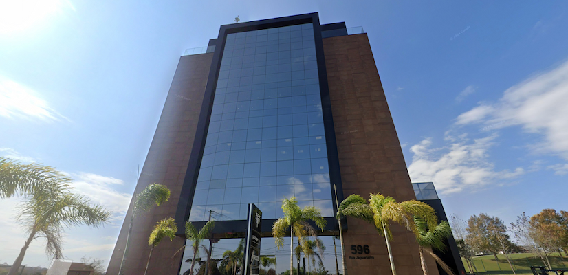 Fachada moderna do edifício-sede do Grupo OM Comunicação Integrada, localizado na Rua Jaguariaíva, 596, em Pinhais (PR). O prédio espelhado de arquitetura imponente reflete o céu azul e as palmeiras do entorno, simbolizando a solidez e a inovação do grupo que abriga a HouseCricket. A imagem transmite profissionalismo e a força do ecossistema criativo do grupo.