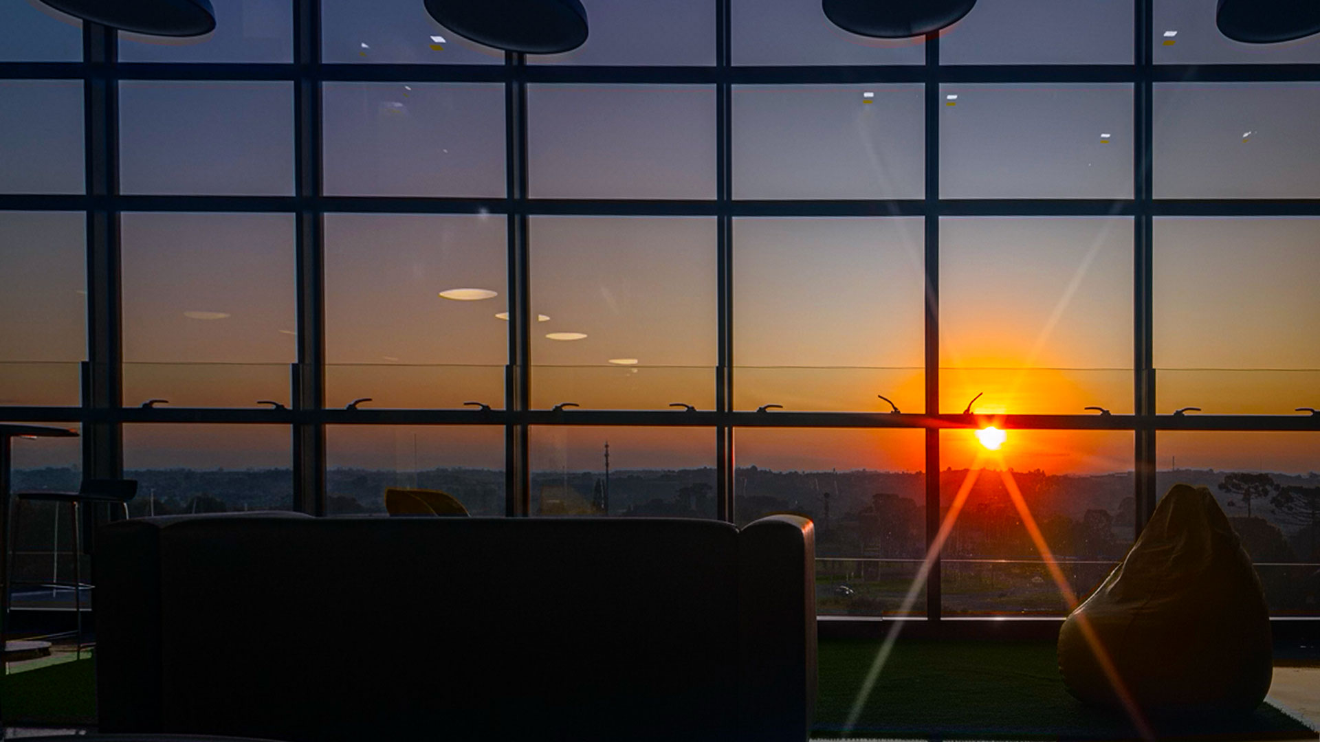 Vista do pôr do sol a partir do interior envidraçado do escritório da HouseCricket, no alto do prédio do Grupo OM, em Curitiba. O sol se põe no horizonte, iluminando o ambiente com luz alaranjada e refletindo nas grandes janelas de vidro. A cena destaca o conforto e a inspiração do espaço de trabalho, marcado por sofás e pufes que simbolizam um ambiente acolhedor, criativo e humano.